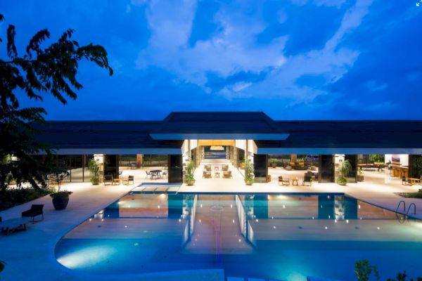 The image shows a large, illuminated pool in front of an open, spacious building under a twilight sky.