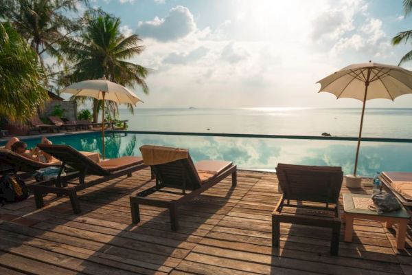 A serene poolside scene with lounge chairs, umbrellas, palm trees, and a view of the ocean under a partly cloudy sky.