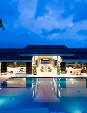 A luxurious, illuminated building at dusk with a large swimming pool in the foreground and surrounding seating areas.