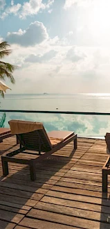A deck with lounge chairs and umbrellas overlooks an infinity pool and ocean with palm trees and a bright sky in the background.