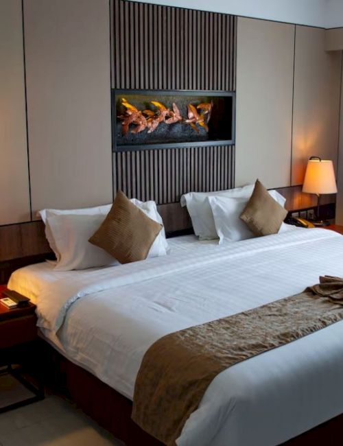 A hotel room features a large bed with brown pillows, side tables with lamps, decorative wall art, and a window with closed curtains.