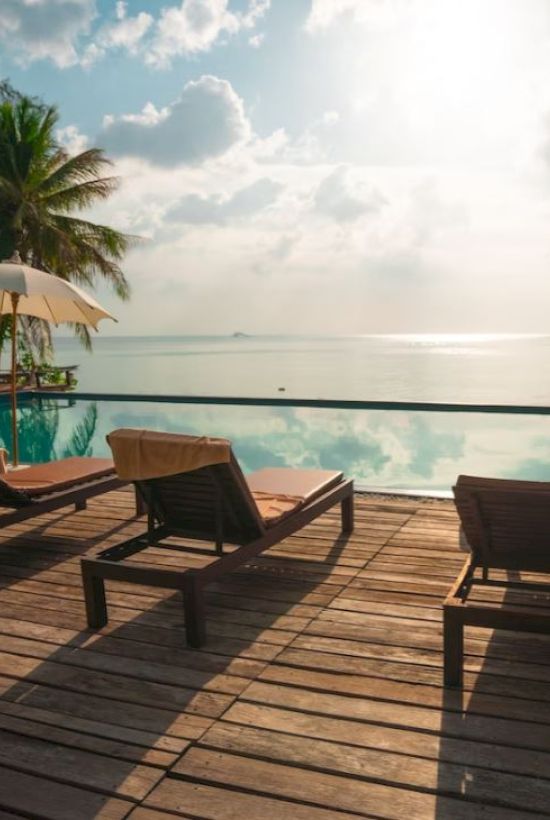 A wooden deck with sun loungers and umbrellas overlooks an infinity pool with a scenic ocean view and palm trees under a bright sky.