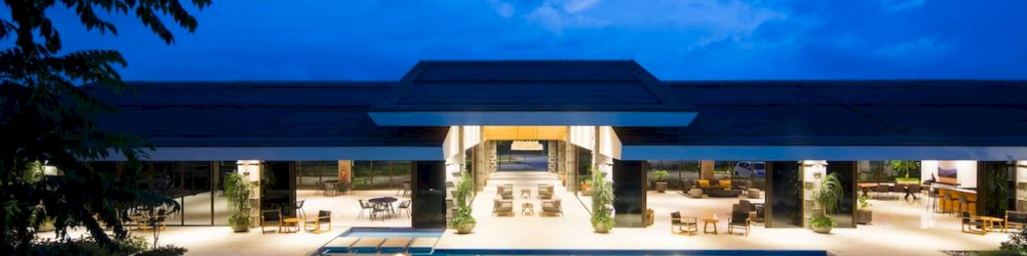 A luxurious, modern building with a brightly lit interior is seen at night, featuring a large outdoor swimming pool reflecting lights.