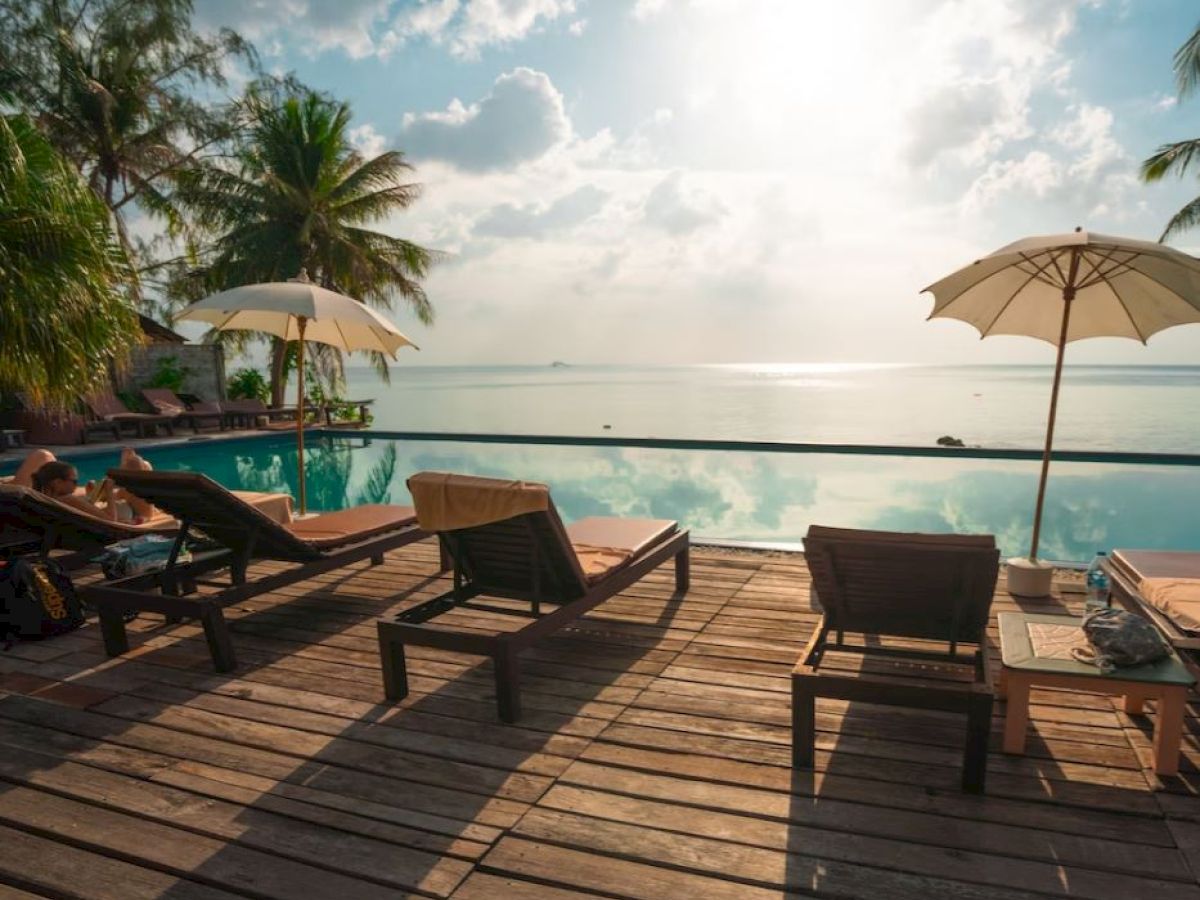 A serene poolside scene with lounge chairs, umbrellas, palm trees, and a view of the ocean horizon under a partly cloudy sky.