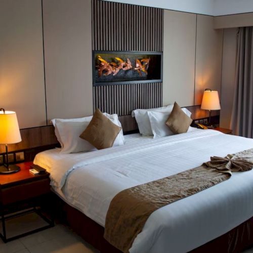 A cozy hotel room features a large bed with pillows, bedside tables with lamps, a wall-mounted artwork, and a window with curtains.