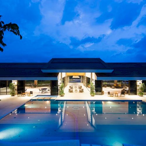A luxurious modern building with a large, illuminated pool in the foreground and a vibrant evening sky above.