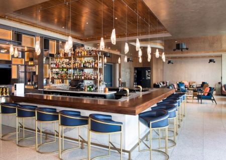 Stylish bar with blue stools, hanging lights, and a well-stocked shelf, set in a modern lounge with cozy seating and wooden accents.