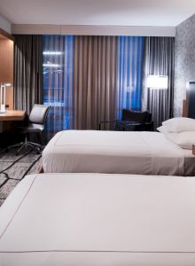 The image shows a modern hotel room with two beds, a TV, a desk, and stylish decor, featuring dim lighting and large windows.