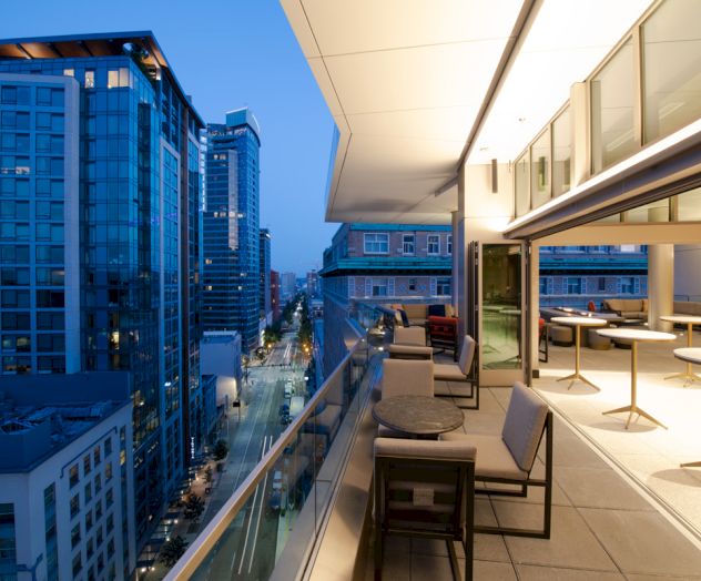 A modern rooftop lounge with seating overlooks a cityscape at dusk, featuring tall buildings and a clear sky.