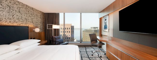 A modern hotel room with a large bed, wall-mounted TV, desk, chair, and a window with a city view, featuring stylish decor.