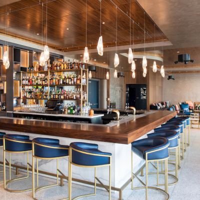 The image shows a modern bar with a wooden countertop, blue bar stools, hanging pendant lights, and a well-stocked back bar area.