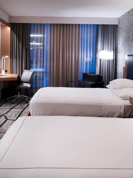 A modern hotel room with two beds, a wall-mounted TV, desk, chair, lamps, and patterned wallpaper creates a cozy atmosphere.