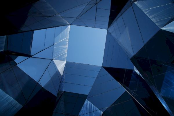 Geometric architecture with reflective glass panels forms an abstract pattern, converging towards a bright blue sky at the center.