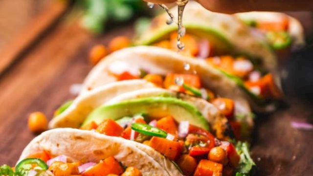 A hand squeezes lime over a row of tacos filled with chickpeas, avocado, and vegetables on a wooden board, garnished with lime slices.
