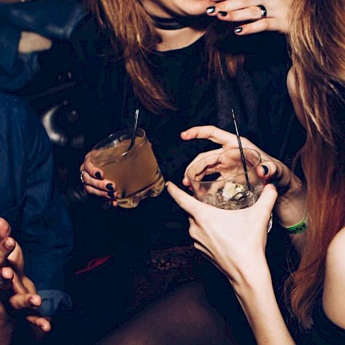 People are socializing and holding drinks in a dimly lit setting, appearing to enjoy a lively conversation.