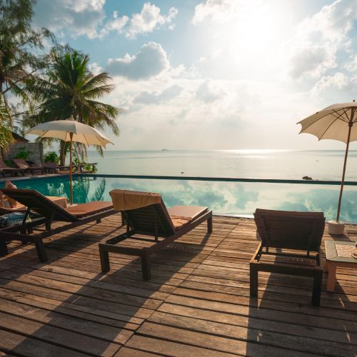 A serene poolside scene with lounge chairs, umbrellas, palm trees, and an ocean view under a partly cloudy sky.