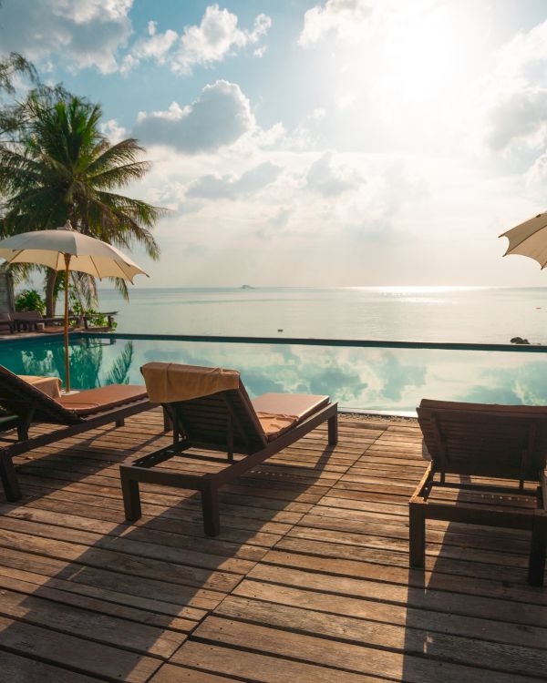 A serene poolside scene with lounge chairs, umbrellas, palm trees, and an ocean view under a partly cloudy sky.