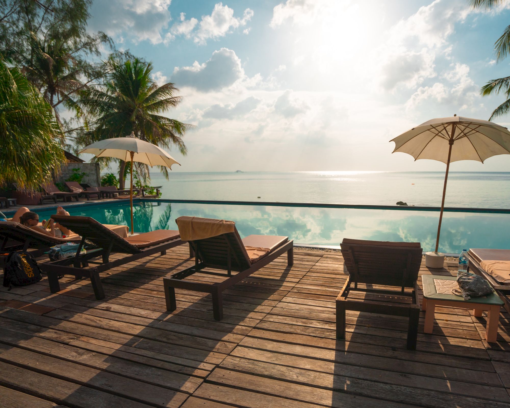 Lounge chairs and umbrellas by a pool overlooking a serene ocean view, with palm trees and clouds under a bright sky.
