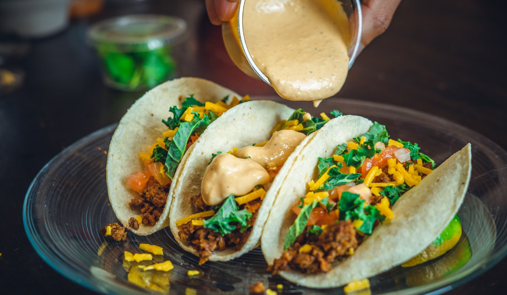 Three tacos with ground filling, veggies, cheese, and sauce being poured on top, served on a glass plate.