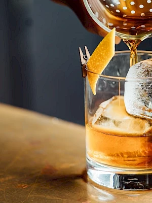 A cocktail being poured over ice in a glass, garnished with a twist of citrus peel, on a bar counter.