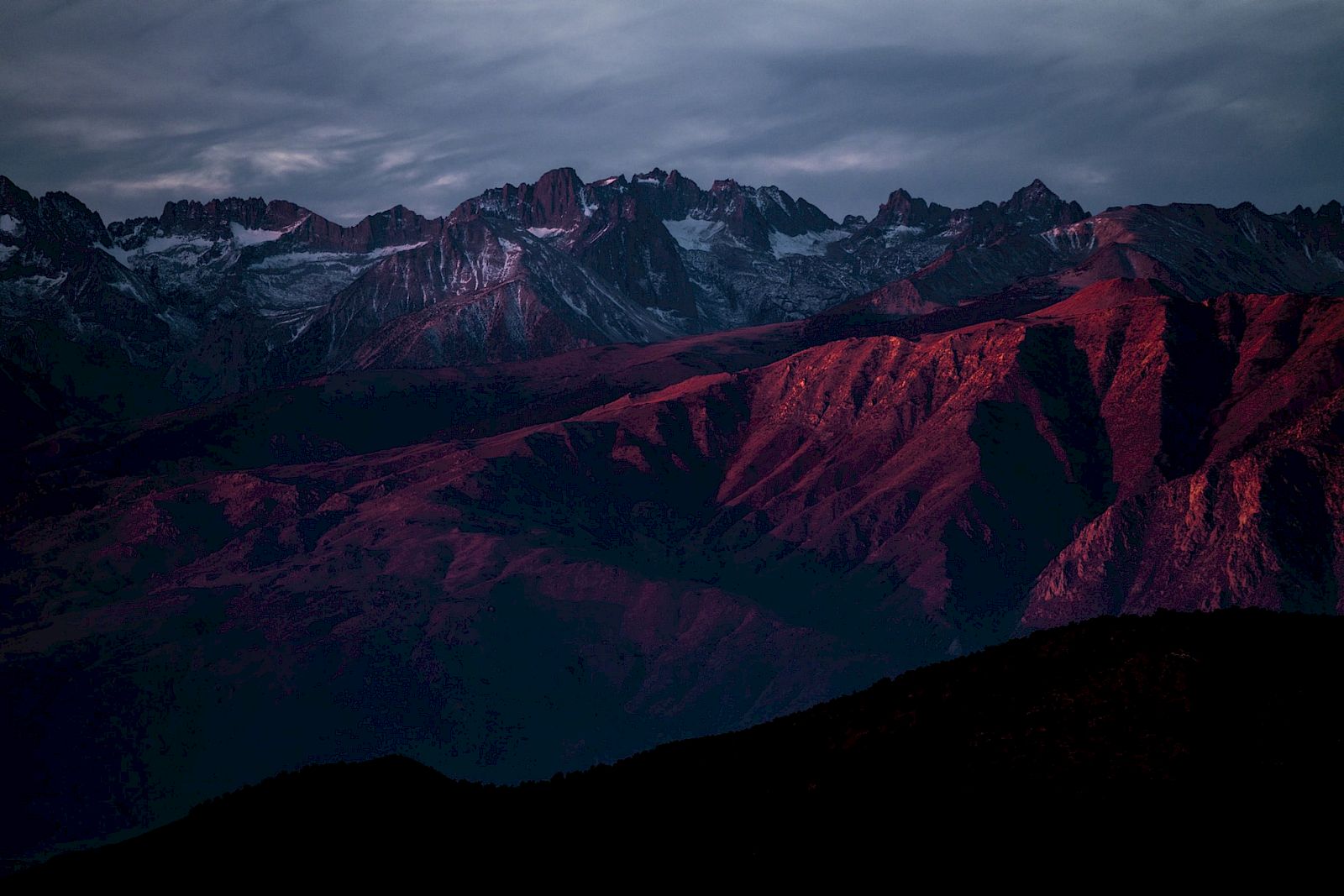 Mountain range with dramatic lighting and snow-capped peaks under a cloudy sky, featuring dark red and purple hues at sunset.