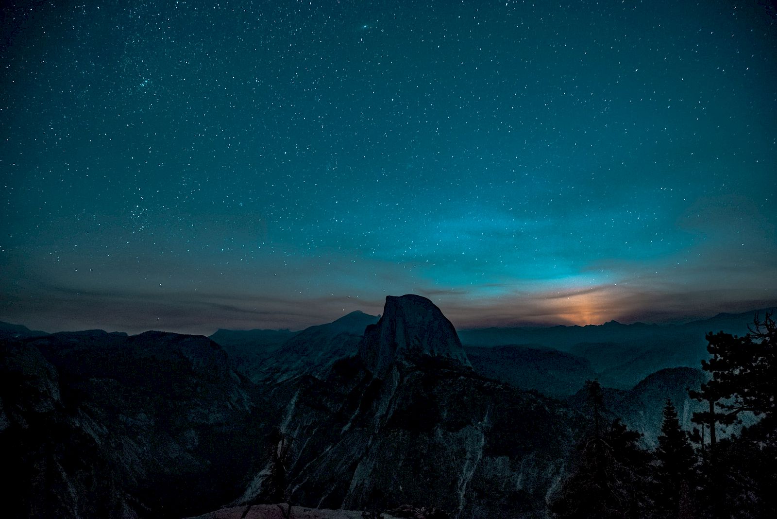 A starry night sky over a mountain range with a glowing horizon and silhouetted trees in the foreground.