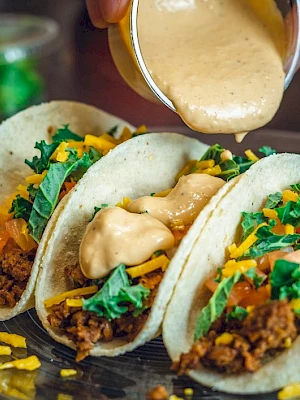 Three tacos filled with ground meat, shredded cheese, lettuce, and tomatoes are being topped with creamy sauce on a plate.