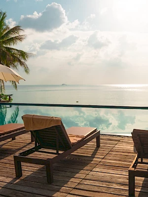 A tranquil poolside scene with lounge chairs, umbrellas, and lush palms overlooks a calm ocean under a sunlit sky.