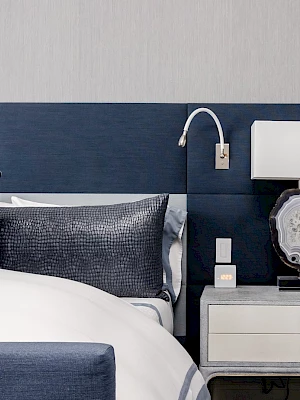 A modern bedroom with a bed, navy headboard, stylish pillows, and a nightstand featuring a lamp and decor. The wall is a light color.
