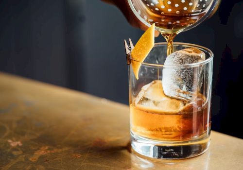 A drink is being poured into a glass with ice and a garnish, likely a cocktail on a bar surface.
