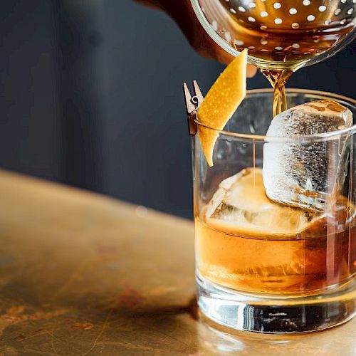 A drink is being poured into a glass with ice and a garnish, likely a cocktail on a bar surface.