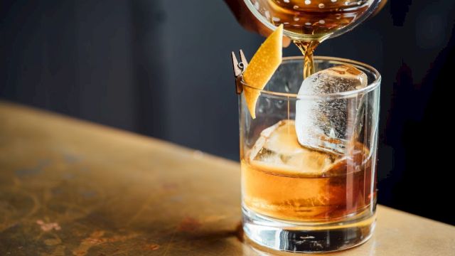 A drink is being poured into a glass with ice and a garnish, likely a cocktail on a bar surface.