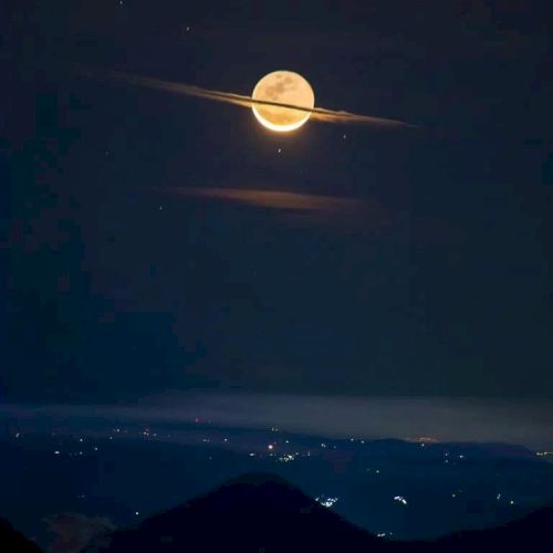 A full moon appears with clouds forming rings around it, resembling Saturn; visible above a landscape with distant city lights.