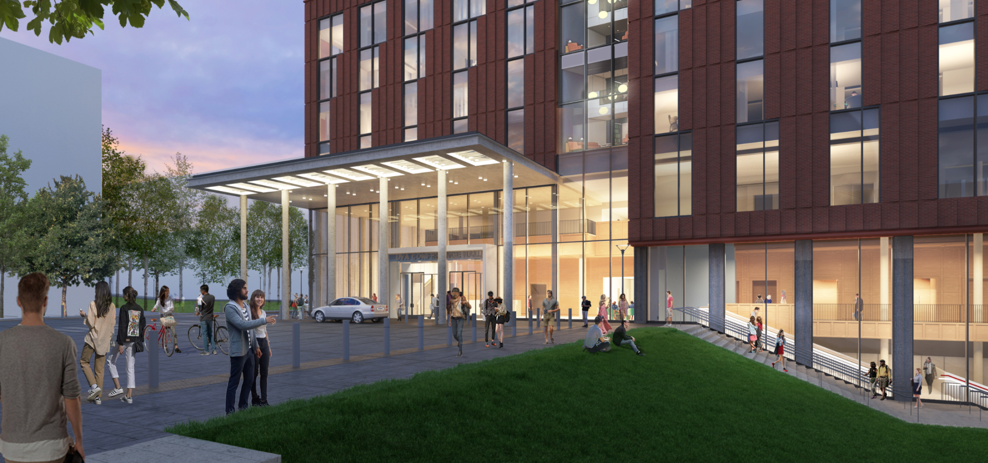 A modern building with a glass entrance and people gathered outside on a grassy area, with bikes and cars nearby.