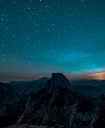 A night sky filled with stars over a mountain landscape, with a glowing horizon suggesting a moonrise or sunset.