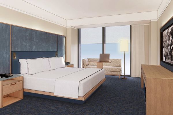 A modern hotel room features a double bed, nightstands, a sofa, a desk lamp, and wall art with large windows providing natural light.
