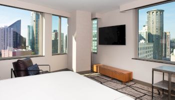 A modern hotel room with a bed, armchair, TV, and large windows offering a city skyline view is shown.