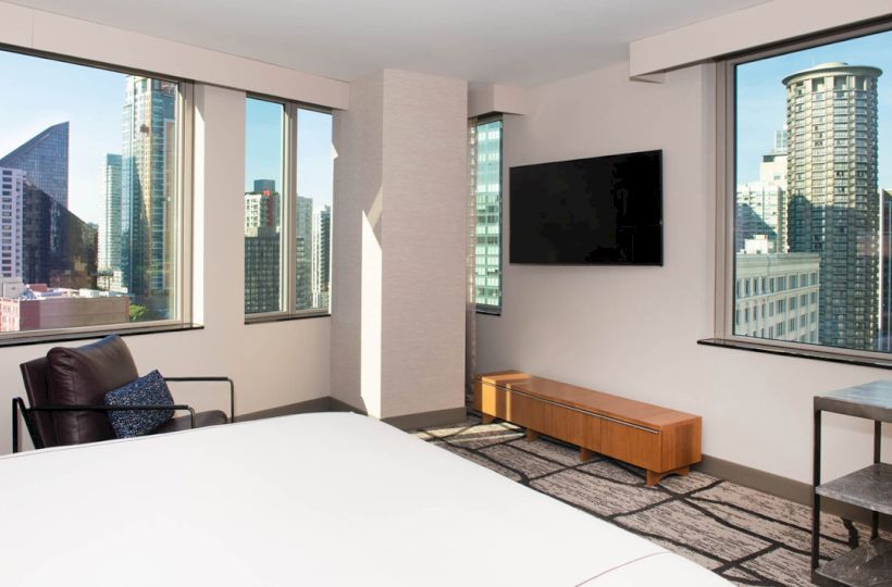 A modern hotel room with a bed, armchair, TV, and large windows offering a city skyline view is shown.