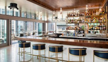The image shows a modern bar interior with a sleek counter, stylish bar stools, and a stocked liquor shelf, set against a cityscape view.