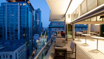 A modern urban balcony overlooking a cityscape at dusk, with seating and tables, surrounded by tall buildings and illuminated windows.