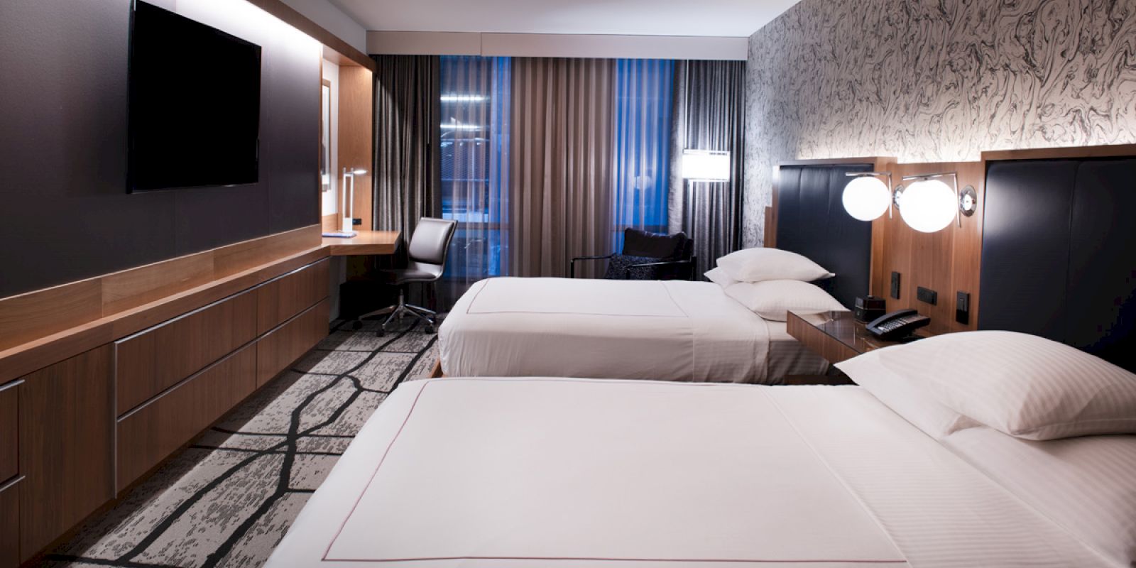 The image shows a modern hotel room with two beds, a TV, a desk, lamps, and patterned curtains and carpet.