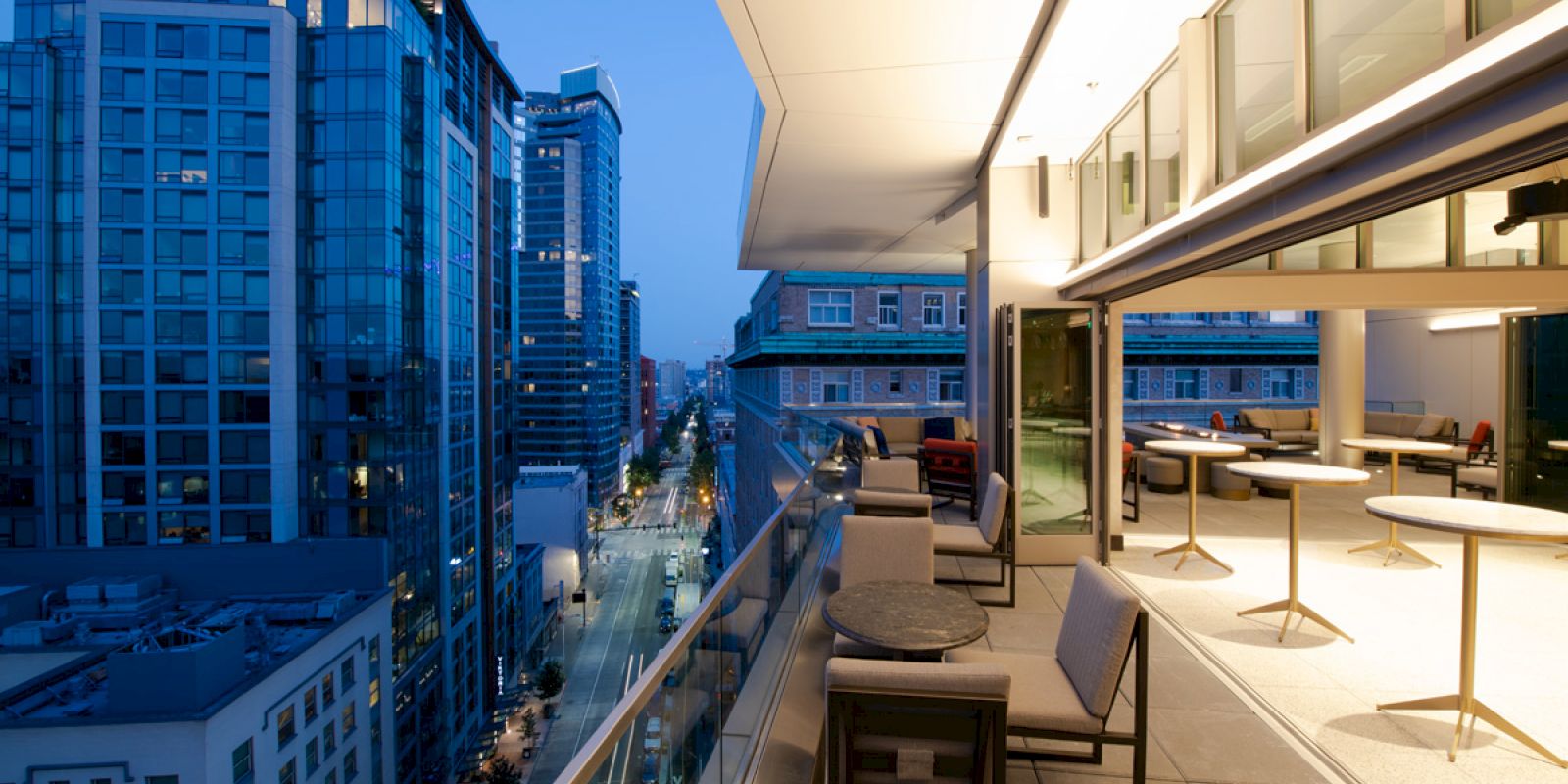 A modern city scene with a rooftop terrace featuring tables and chairs, overlooking a cityscape with tall buildings during twilight.