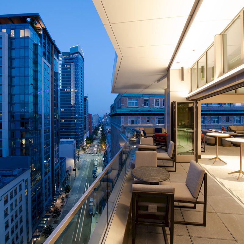 A modern balcony view on a high-rise building overlooking a city street, with tables and chairs, at dusk.
