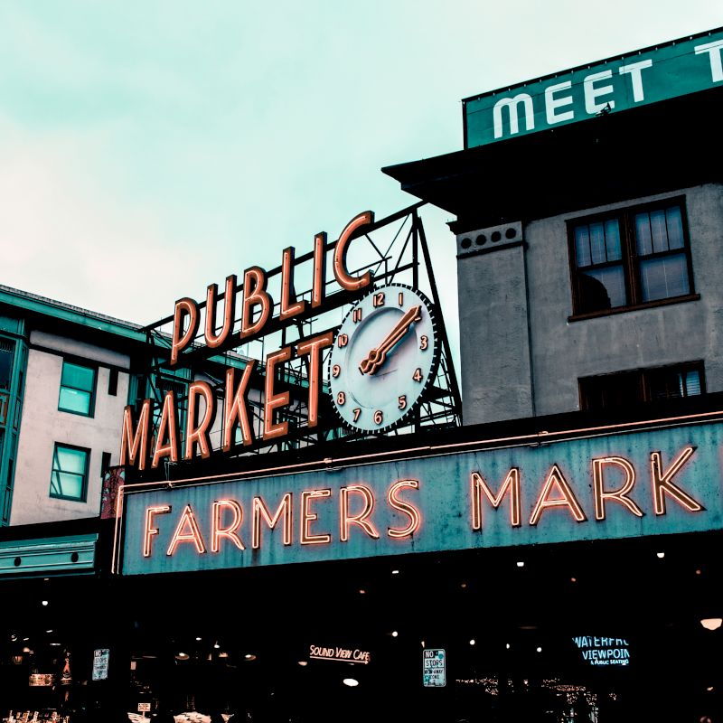 The image shows a public market sign with a clock, reading "Farmers Market," in front of historic buildings with a vintage appearance.