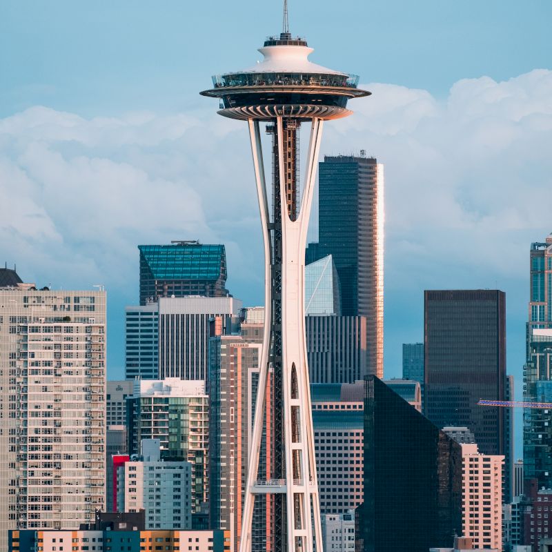 The image shows the Space Needle in Seattle, with a backdrop of modern skyscrapers under a partly cloudy sky.