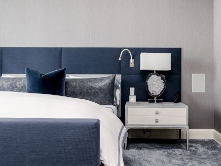 A modern bedroom with a blue upholstered bed, white bedding, blue pillows, a nightstand with a lamp, and decorative accents.