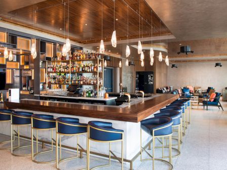 A modern bar with a central counter, stylish blue barstools, a well-stocked liquor display, hanging lights, and a cozy seating area.