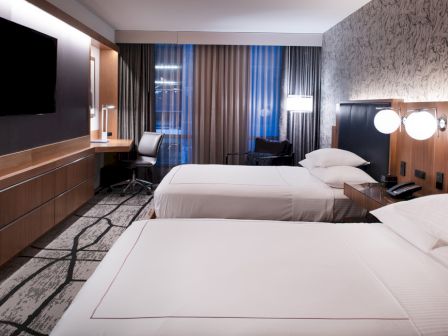 The image shows a modern hotel room with two beds, a TV, desk, lamps, and patterned carpet, featuring a mix of dark and light decor.