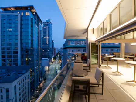 A modern cityscape at dusk with high-rise buildings viewed from a stylish balcony featuring tables and seating.