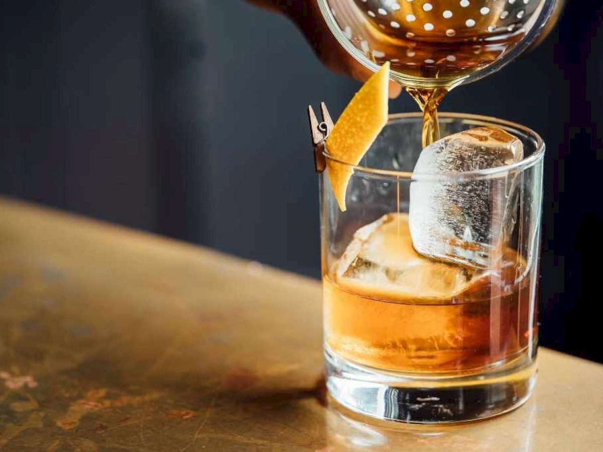A cocktail being poured into a glass with ice and a citrus peel garnish, resting on a bar counter.
